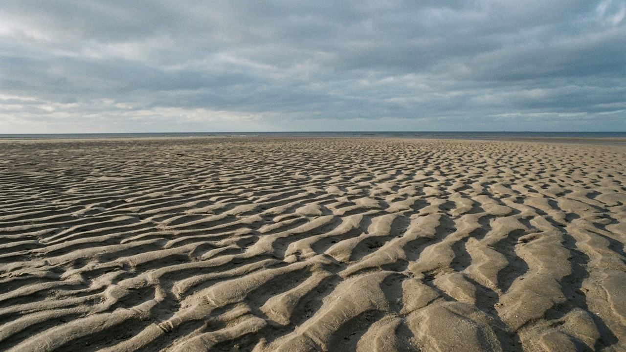 Overlapping ridges of wind blown sand on an empty beach at low tide, long shadows, wide grey sky